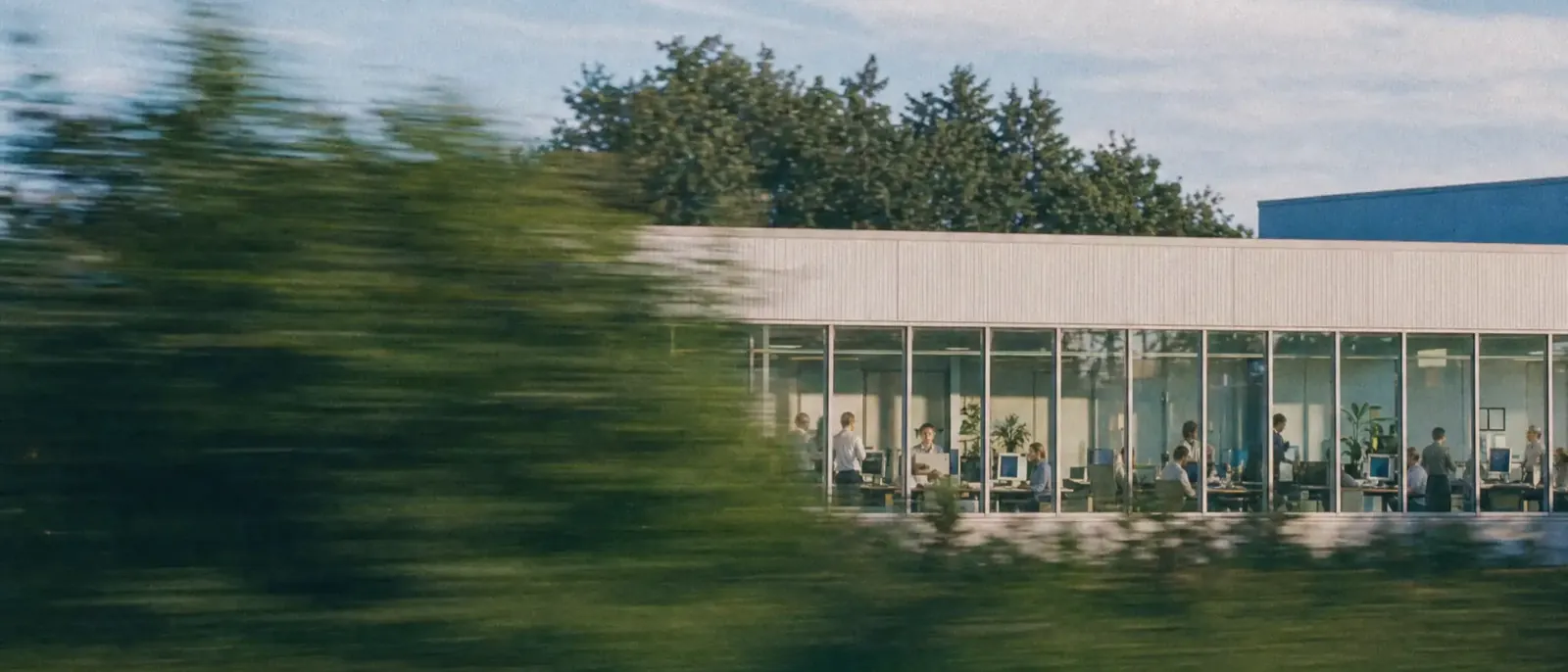 A glass office seen from a moving train, people at their desks inside, a summer sky above