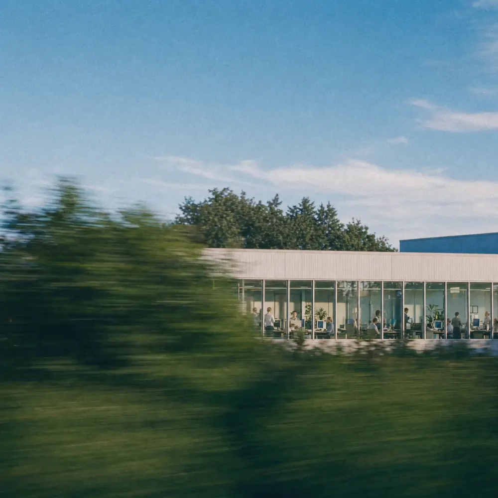 A glass office seen from a moving train, people at their desks inside, a summer sky above